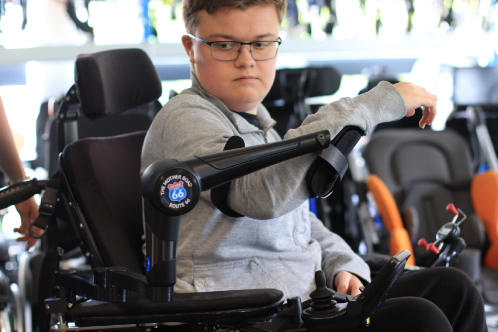 You see an ORTHOPUS Supporter robotic arm attached to a person’s electric wheelchair. The ORTHOPUS Supporter is customized with an image and colorful shells in different shades.