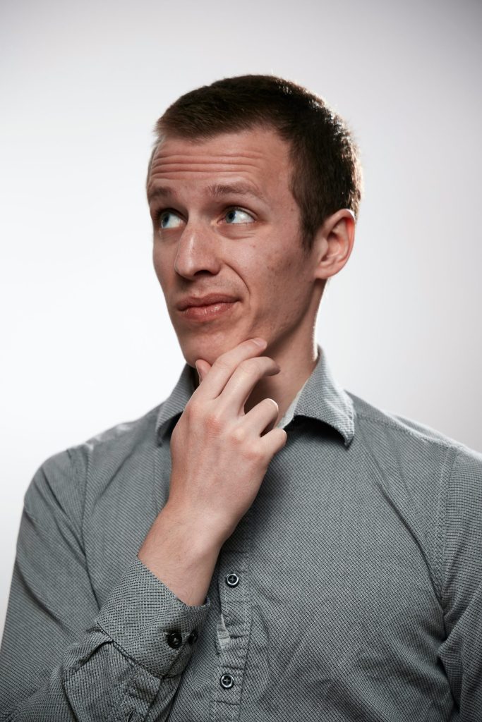 Photo d'un homme sur fond blanc. L'homme a les cheveux court et les yeux bleus, il porte une chemise grise et mime de se poser une question.
