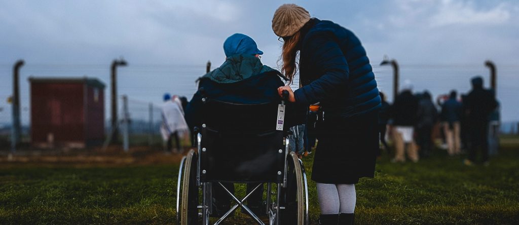 Photo d'une personne en fauteuil roulant avec une autre personne à côté d'elle qui s'abaisse à sa hauteur, ils regardent tous les deux l'horizon, ils ont une vue sur un champ et une sorte de cabane en bois sur la gauche.