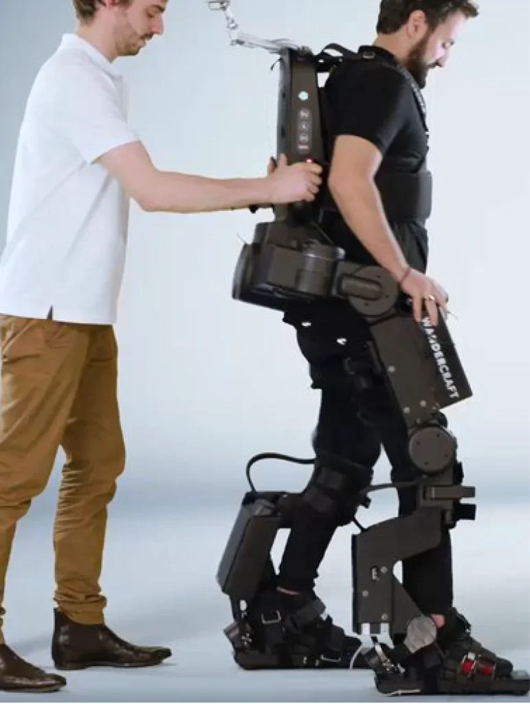 Un homme en polo blanc et chino camel est derrière un autre homme qui a un exosquelette médical du bas des jambes jusqu'en haut de de sa nuque.