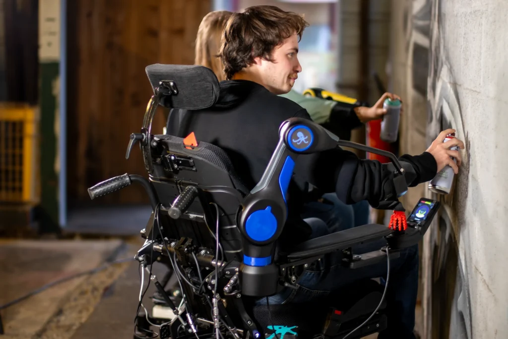 Jeune homme utilisateur de l'ORTHOPUS Partner qui grâce à cet assistant robotique qui maintient son bras, taggue sur un mur. L'assistant robotique est bleu et noir branché sur son fauteuil roulant électrique noir.