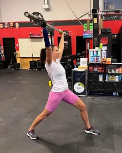 Femme qui soulève des haltères grâce à une prothèse à l'un de ses bras. Elle est dans une salle de sport, sa prothèse est noire et elle porte un débardeur blanc ainsi qu'un leggings rose.