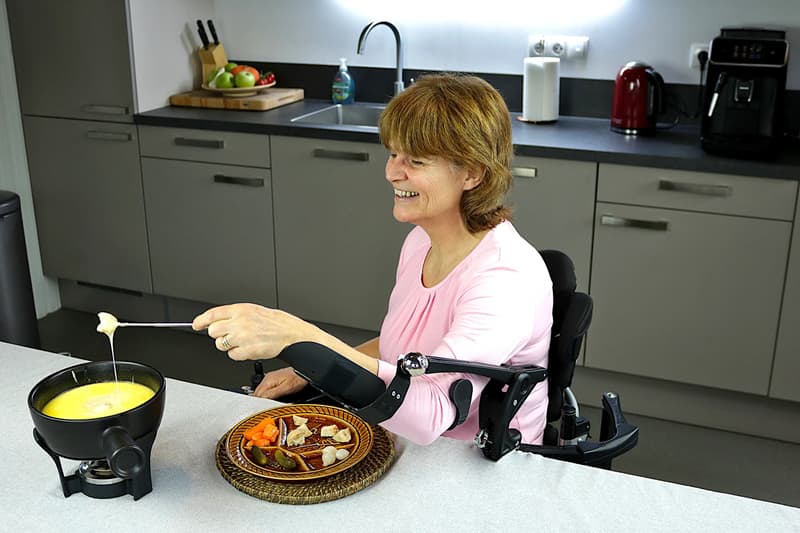 Photo d'une femme qui, grâce à un bras mécanique et une orthèse, trempe un morceaux de pain dans du fromage pour une fondue. Devant elle il y a une assiette et l'appareil à fondue. La femme a environ une soixantaine d'année et est châtain clair avec une frange, elle a également un pull rose et elle est dans sa cuisine.