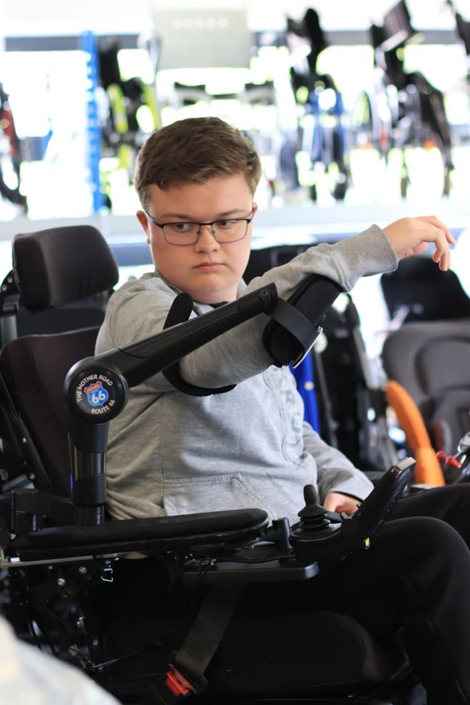 Jeune homme avec des lunettes. Il est dans un fauteuil roulant électrique et il lève le bras grâce à un robot, l'ORTHOPUS Supporter.