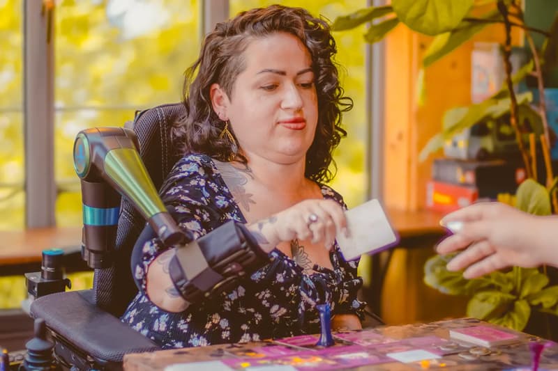 Une jeune femme en fauteuil roulant en train de jouer à un jeu de société. Elle lève le bras pour donner une carte à quelqu'un grâce à un assistant robotique pour la mobilité des bras, l'ORTHOPUS Supporter.