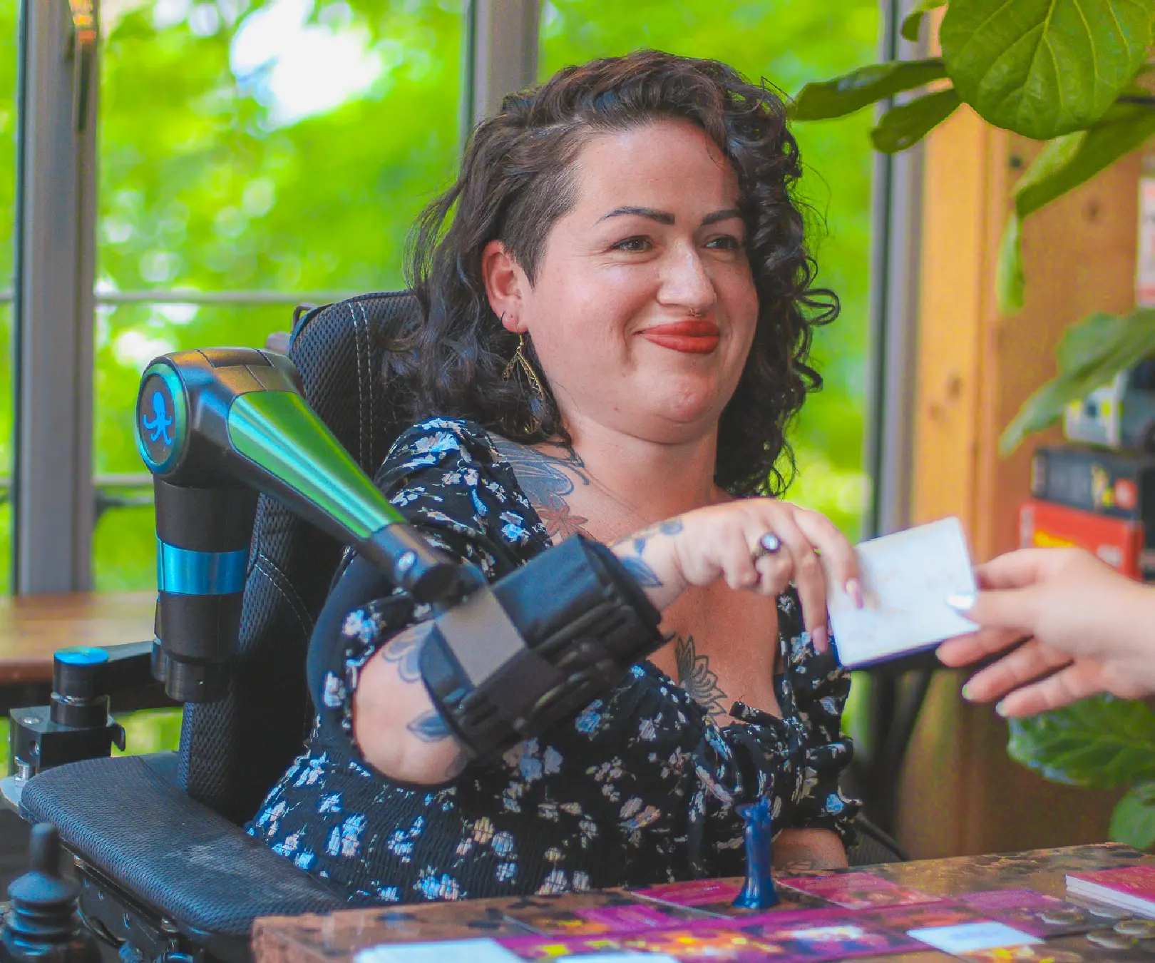 Smiling woman in a wheelchair using a robotic arm to hand a card to someone, seated at a table in a bright room with plants in the background.
