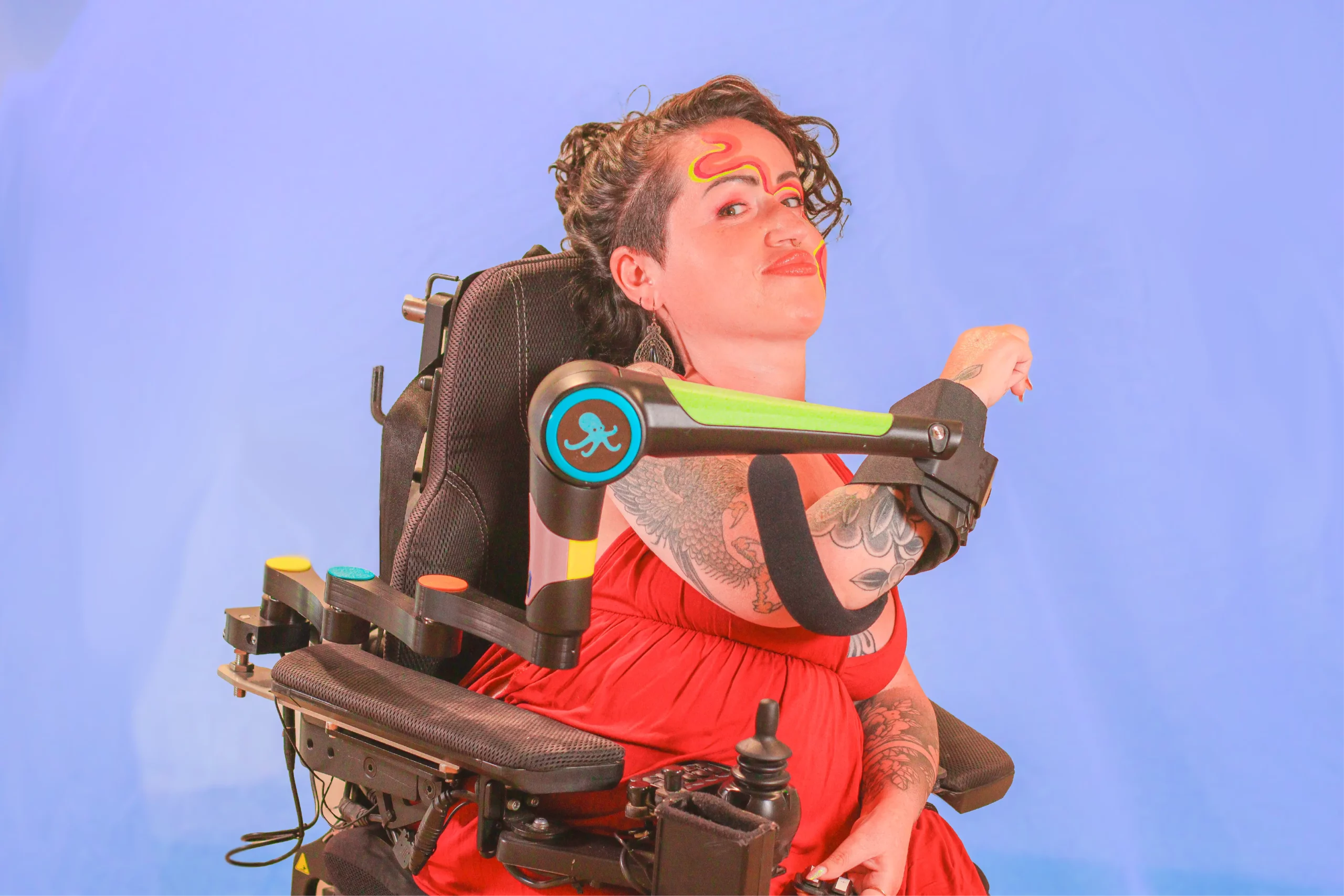 A woman in a wheelchair wears a robotic assistive device, the ORTHOPUS Supporter, to aid arm mobility and poses in front of a blue background.