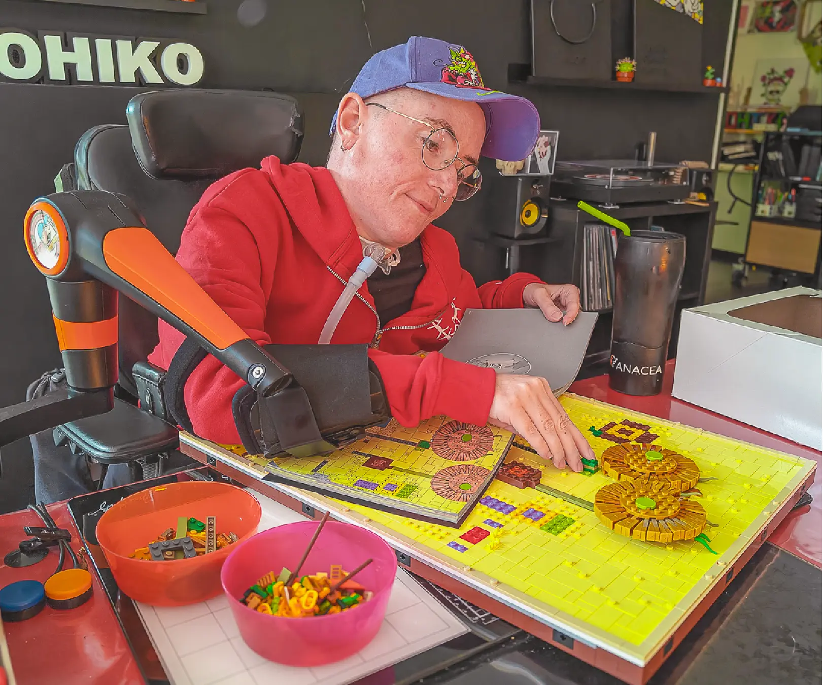 A person wearing a blue cap and a red sweatshirt, seated in a wheelchair equipped with the orange and black robotic assistive device, the ORTHOPUS Supporter, assembling a colorful LEGO mosaic on a large yellow baseplate, surrounded by bowls of LEGO pieces, in a personalized living space.